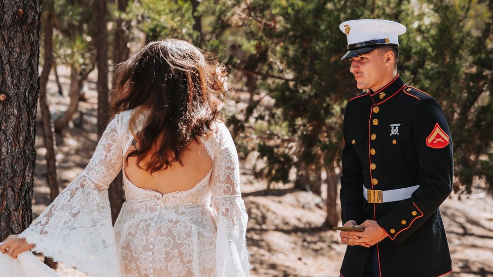 A Marine in dress blues shares a first look moment with a bride in a white lace gown among pine trees.
