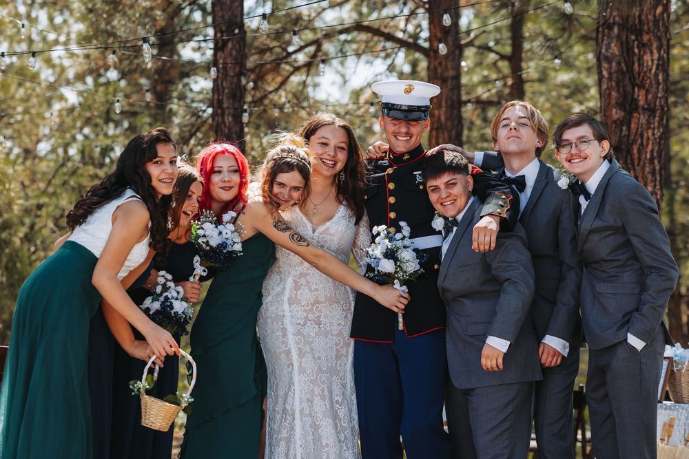 Wedding party posing outdoors in forest setting, bride in lace gown with groom in Marine dress uniform and guests in green.
