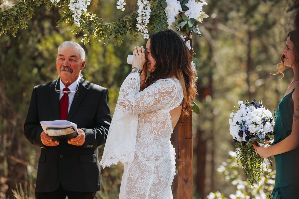 Emotional bride in lace wedding dress wiping tears during outdoor forest ceremony with officiant and bridesmaid.