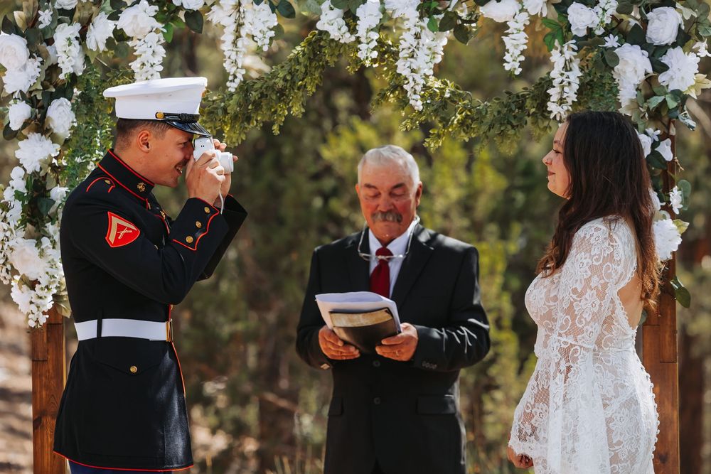 Marine groom wipes tears at outdoor wedding ceremony under floral arch with bride and officiant.