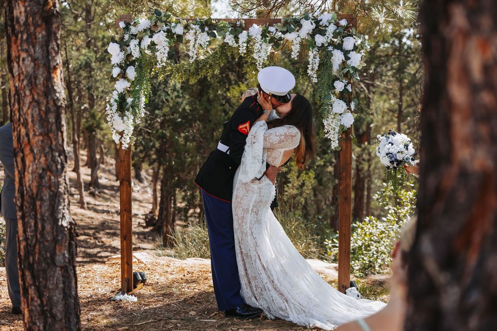 Military groom in dress uniform kisses bride in lace gown under floral arch in pine forest wedding ceremony.