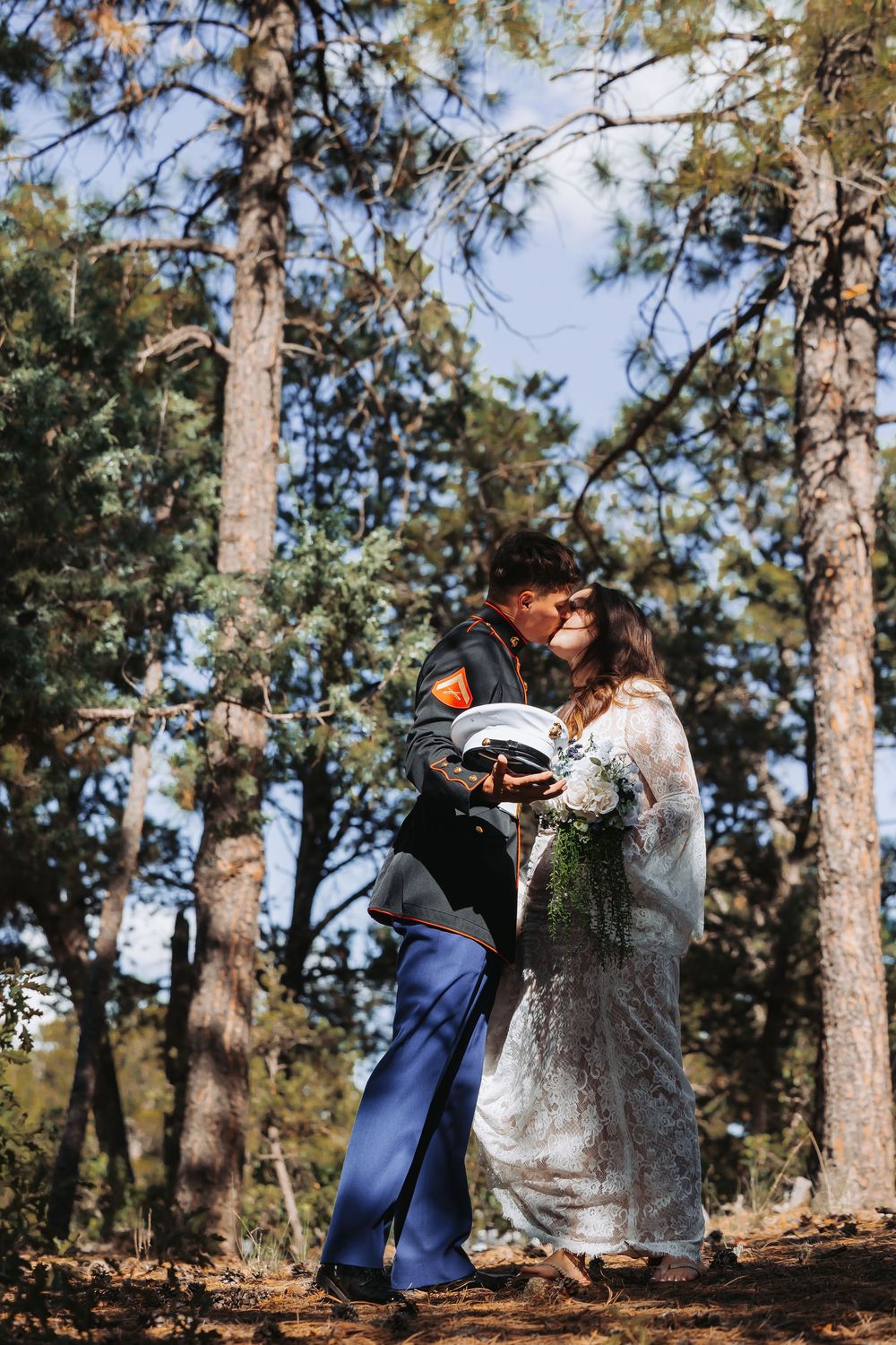 Military groom and bride in lace dress share a kiss in a pine forest, bride holding a white floral bouquet.