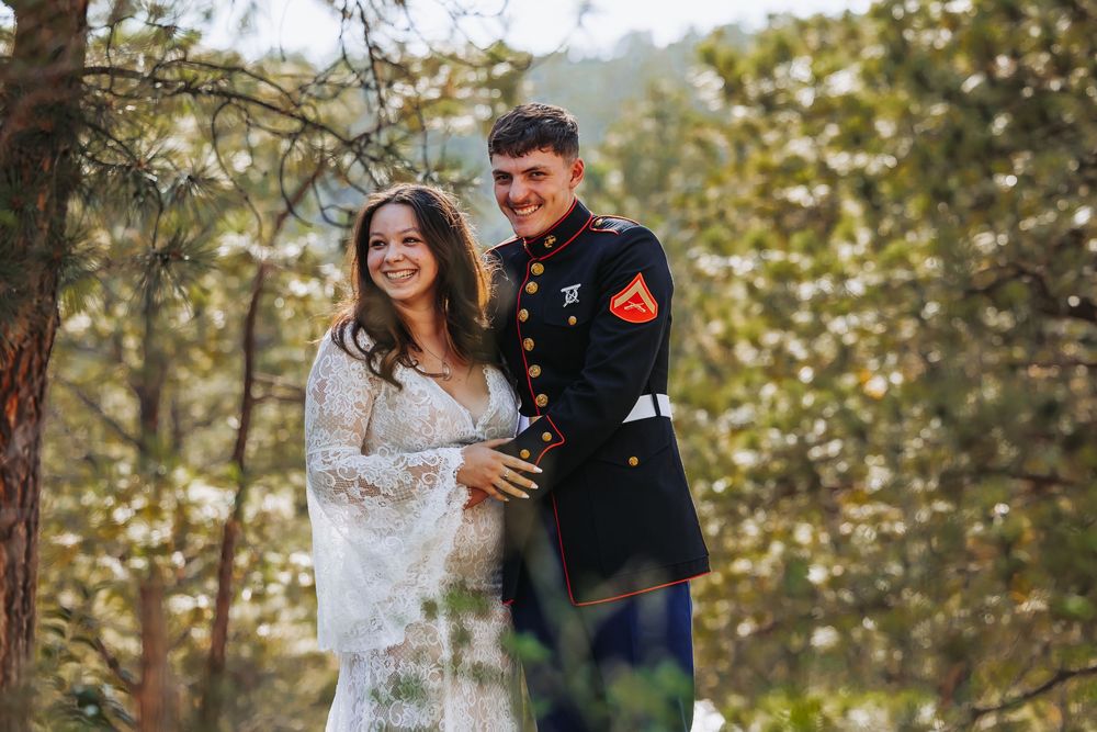 Pregnant woman in lace dress with Marine in dress blues smiling outdoors among trees.