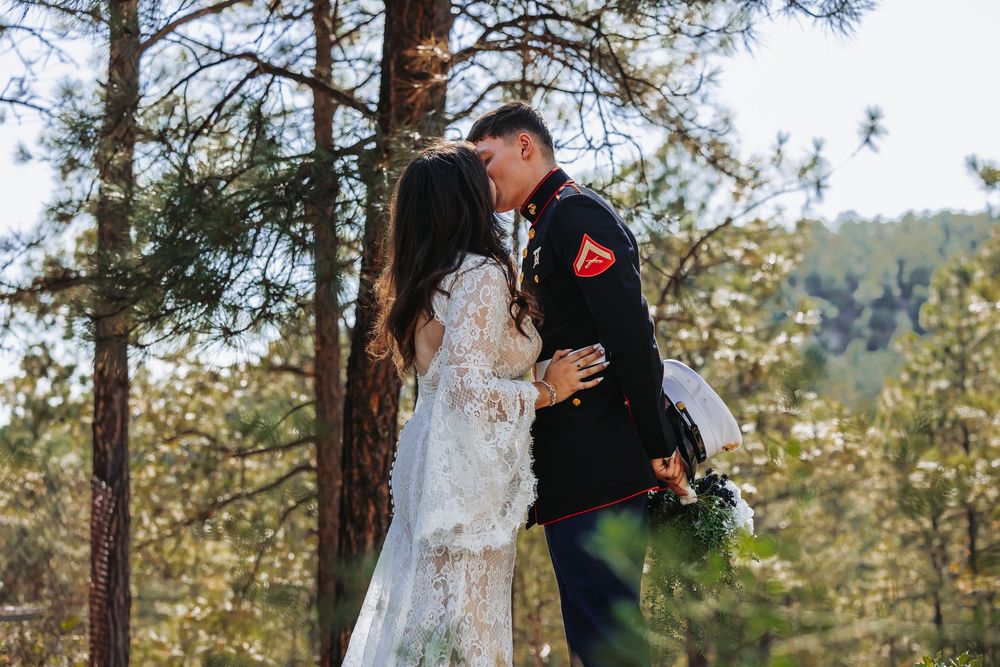 A Marine in dress uniform shares a romantic kiss with his bride in a white lace gown among pine trees.