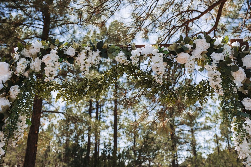 Floral wedding arch adorned with white roses and greenery set among tall pine trees outdoors.