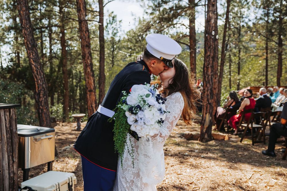 Marine in dress uniform kisses bride holding white bouquet at outdoor forest wedding ceremony with guests seated behind.