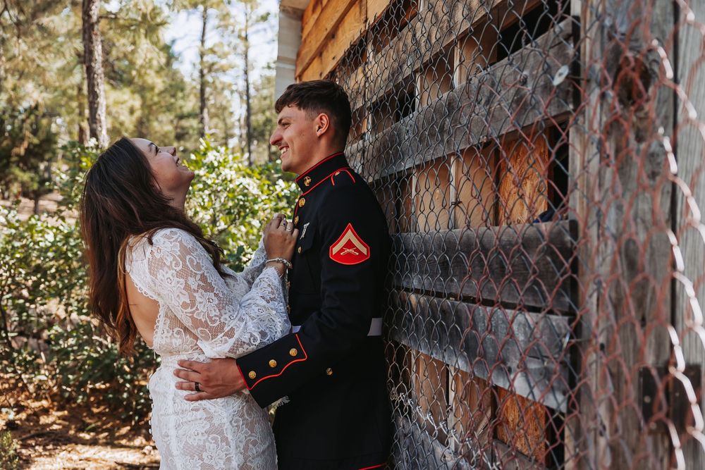 A Marine in dress uniform embraces his bride in a white lace gown outdoors near a rustic fence.