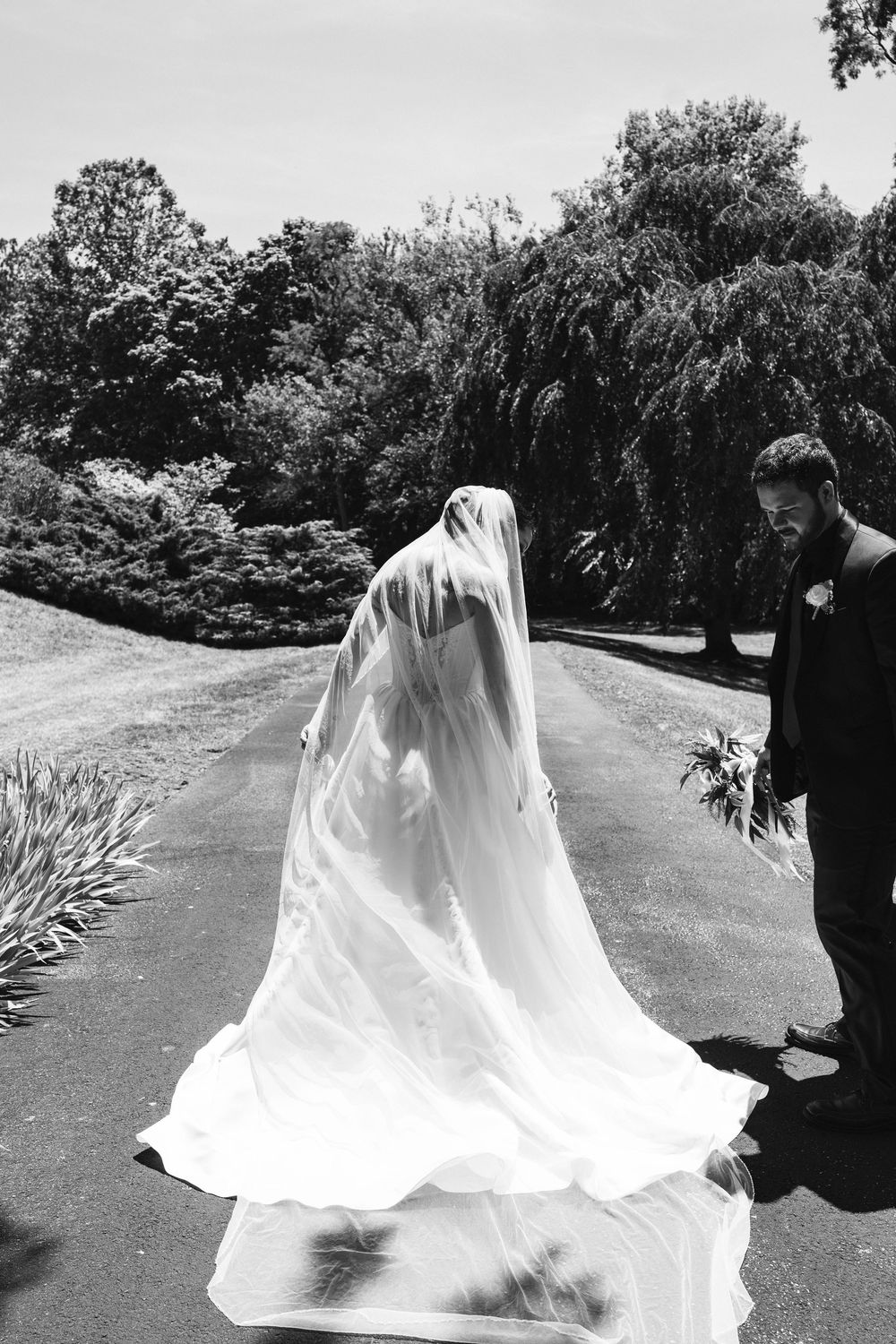 Black and white photo of a bride in a flowing veil and gown walking with a groom on a tree-lined path.