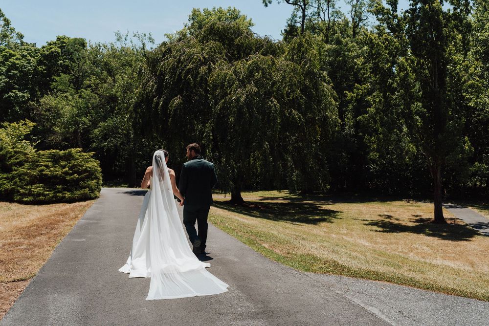 Bride and groom walking hand in hand down a tree-lined path, bride wearing a long white gown with flowing veil.