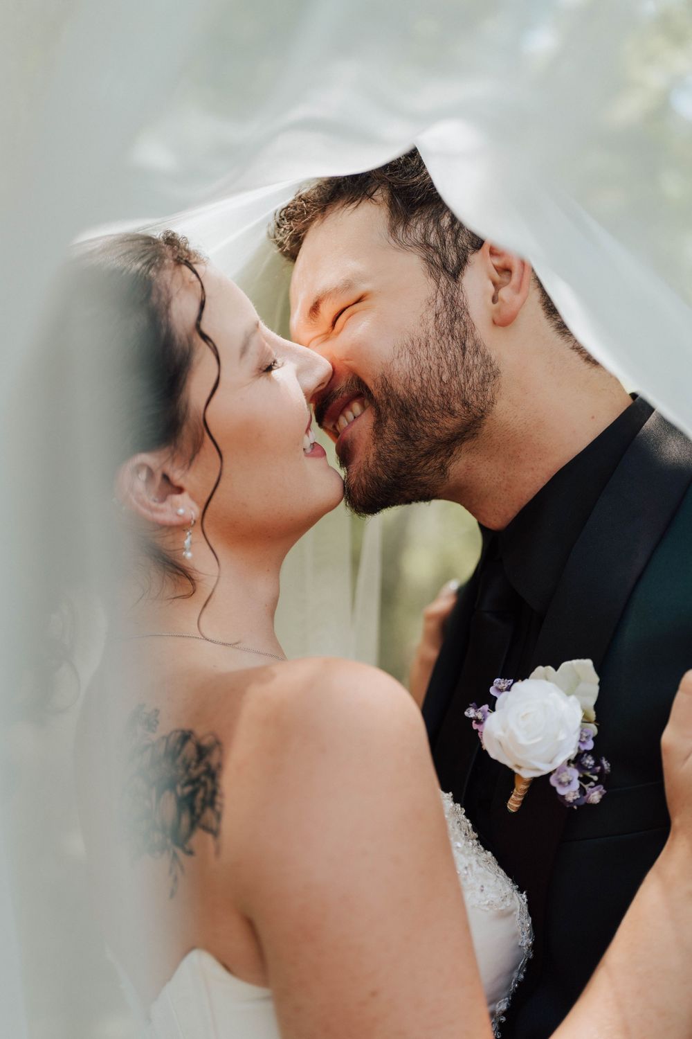 Bride and groom sharing romantic moment under wedding veil, groom in black suit with white boutonniere, bride with tattoo.
