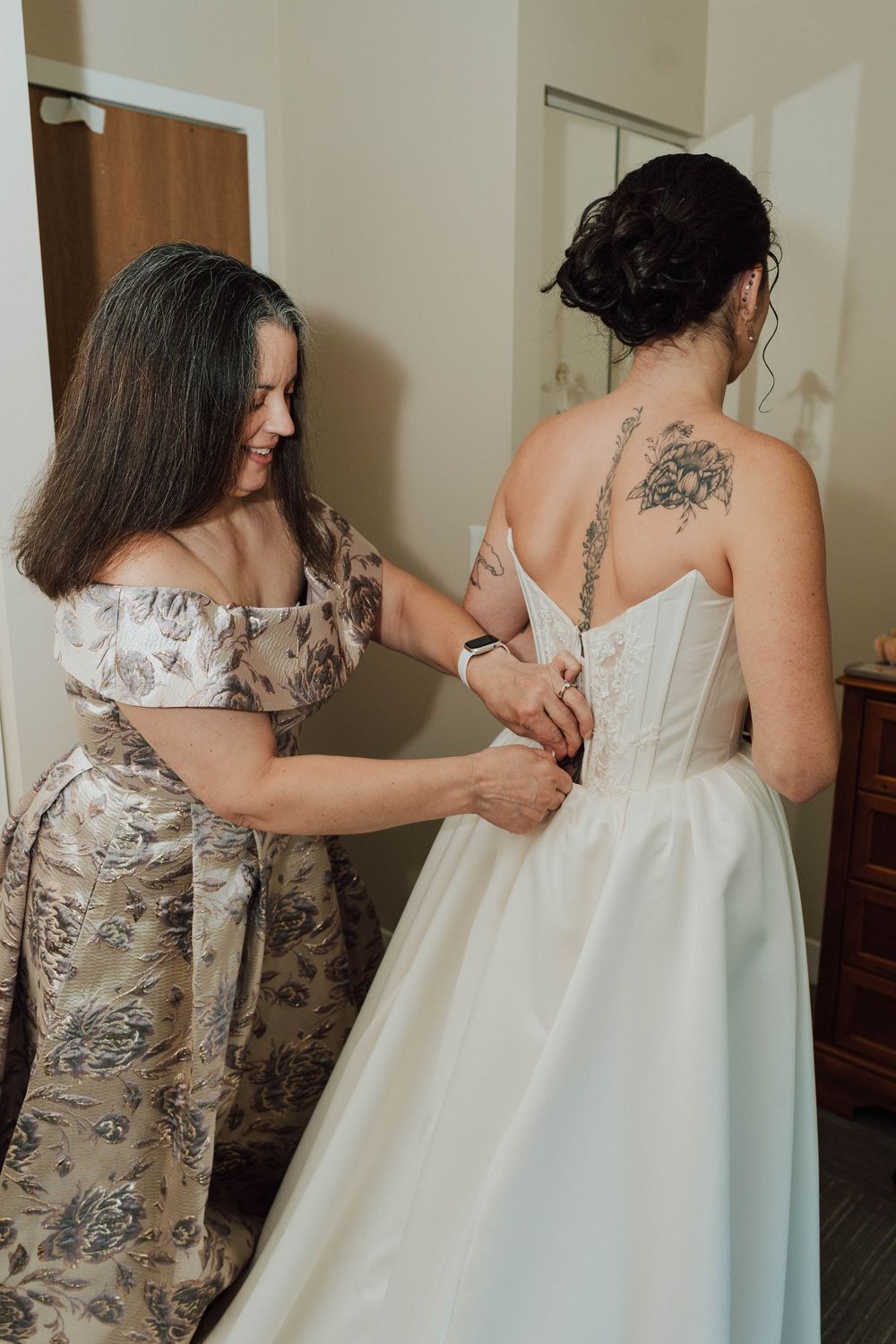 A woman helps a tattooed bride fasten her white corset wedding dress from behind in a bridal suite.