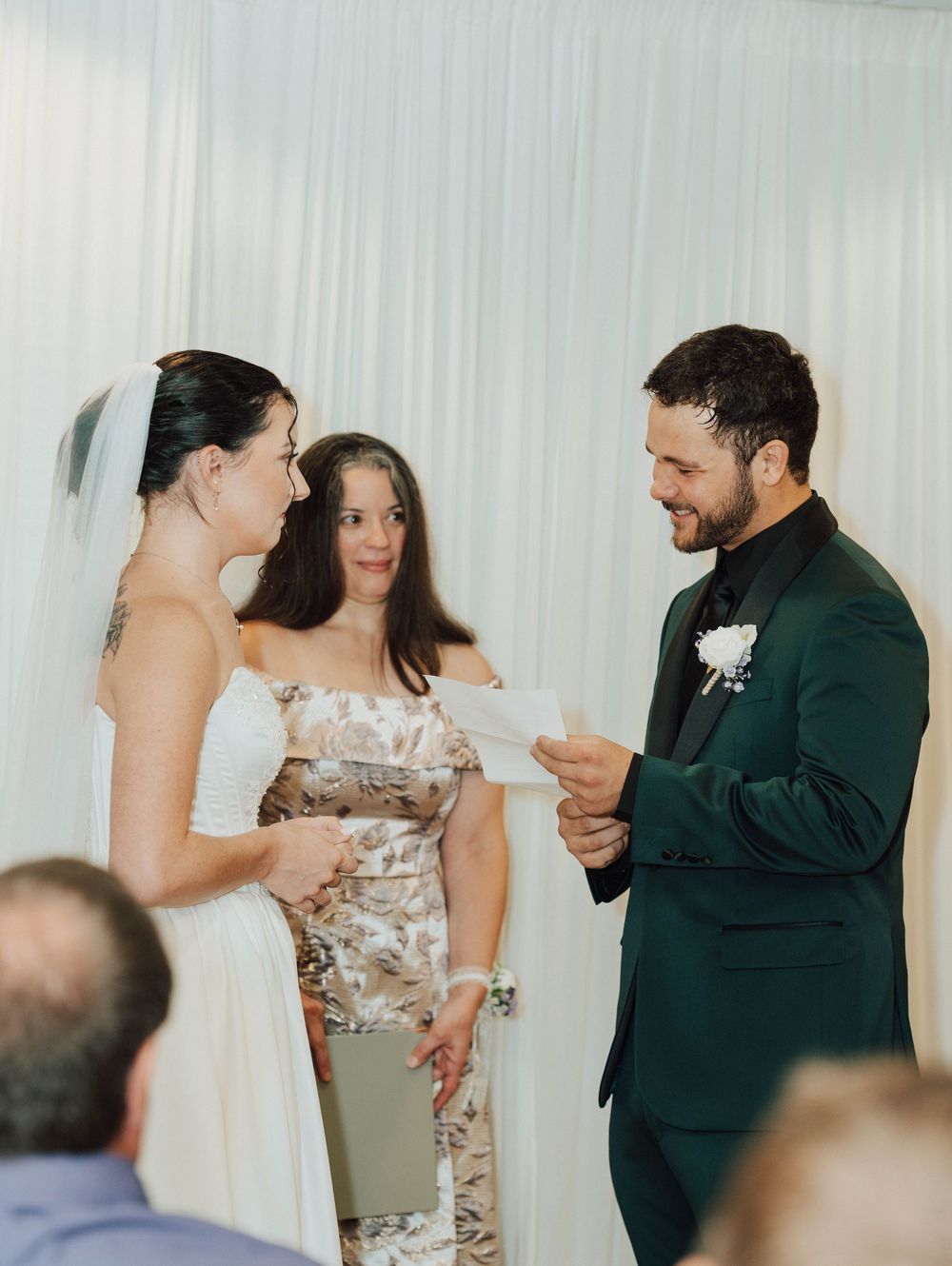 Groom in green suit reads vows to bride in white dress during wedding ceremony with officiant behind them.