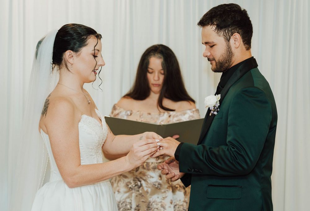 Bride placing wedding ring on groom's finger during ceremony, officiant holding book in background.
