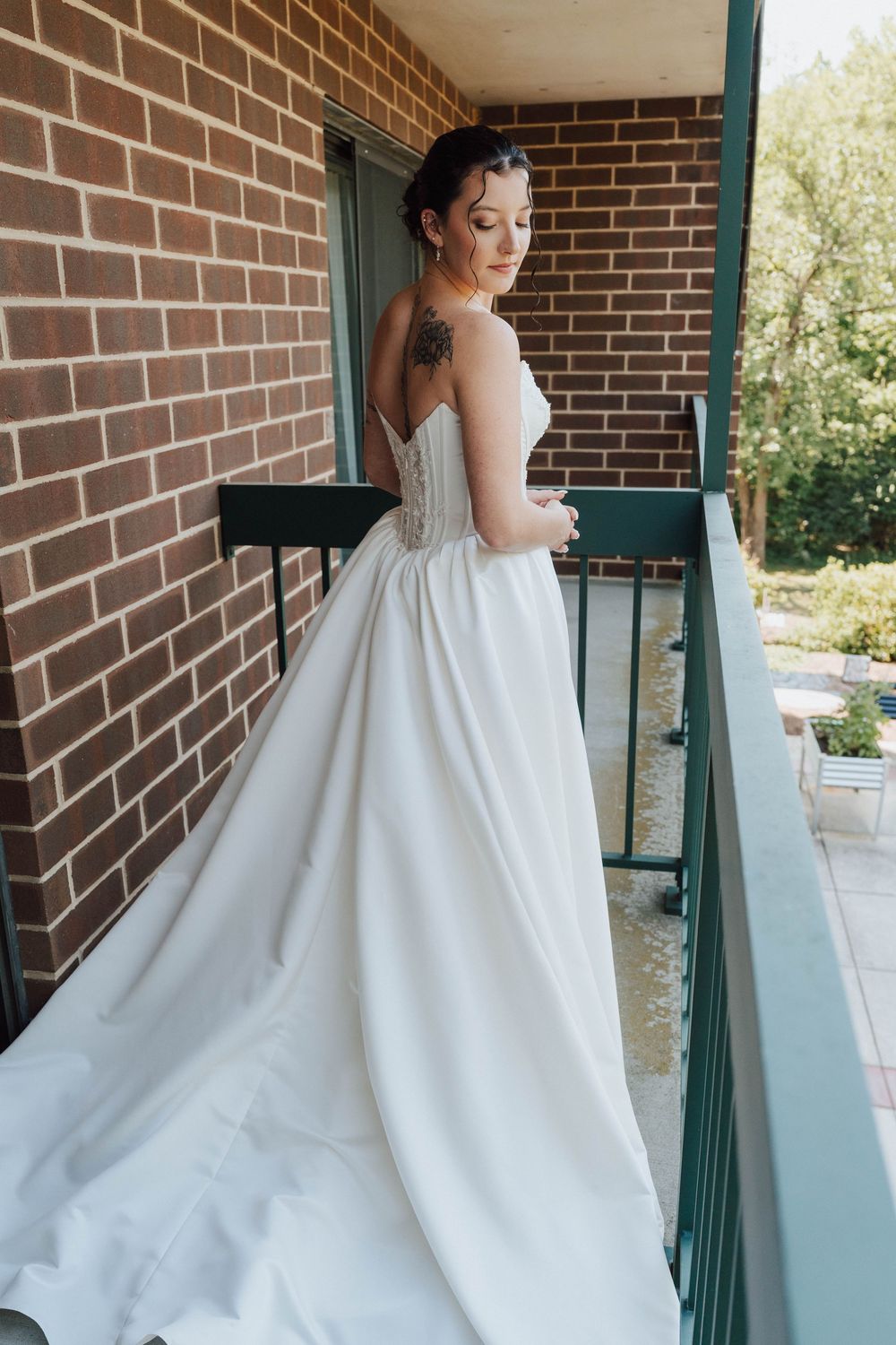 Bride in elegant backless white wedding gown with corset detail stands on brick balcony, showcasing long flowing skirt.