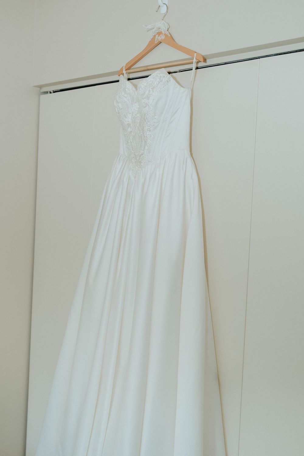 White A-line wedding dress with lace bodice and spaghetti straps hanging on wooden hanger against closet doors.