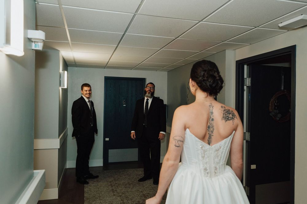 Bride in white gown with back tattoo faces two groomsmen in black suits in a hotel hallway during wedding first look.