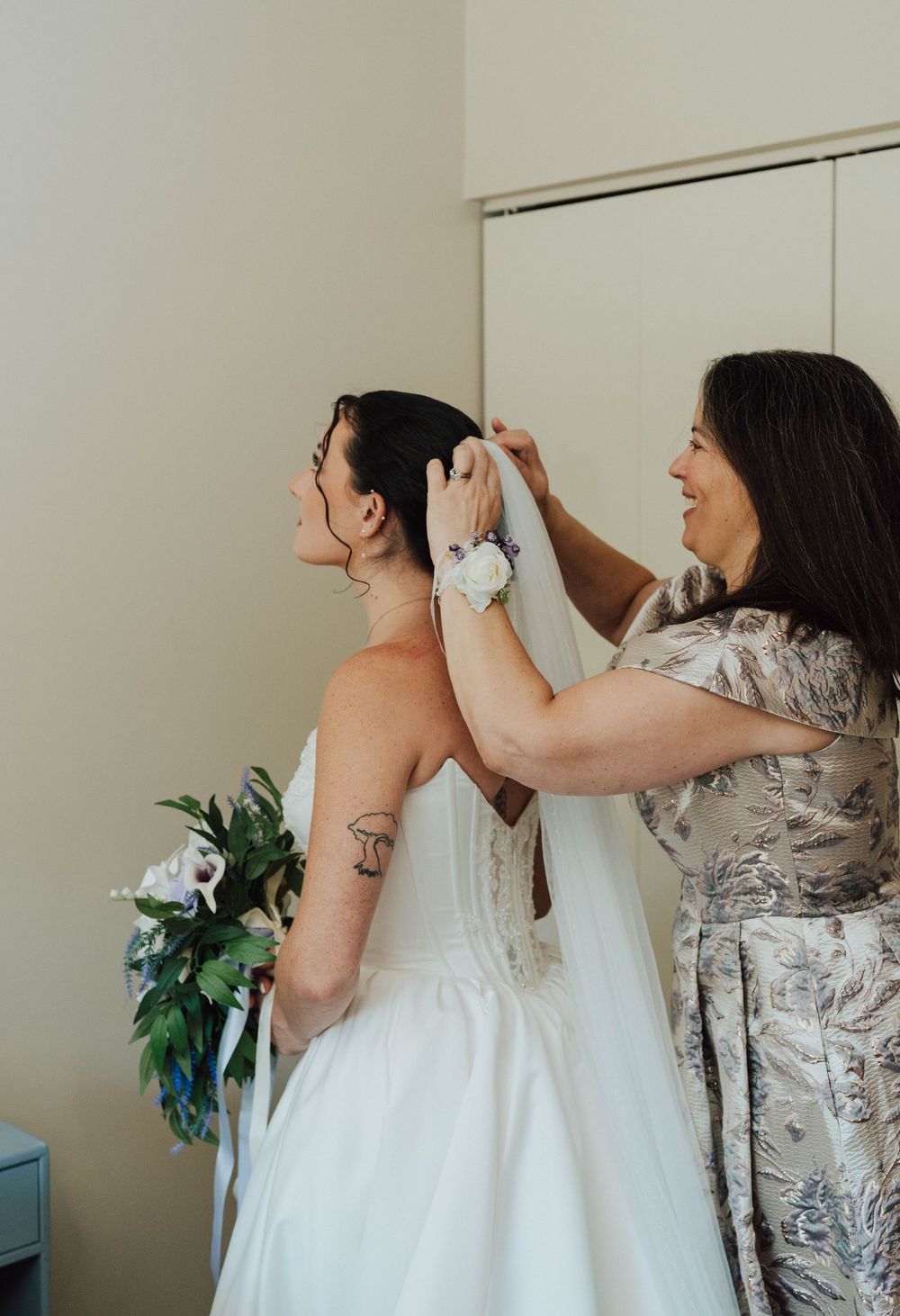 A woman in a white wedding gown holds a bouquet while another woman helps adjust her veil before the ceremony.