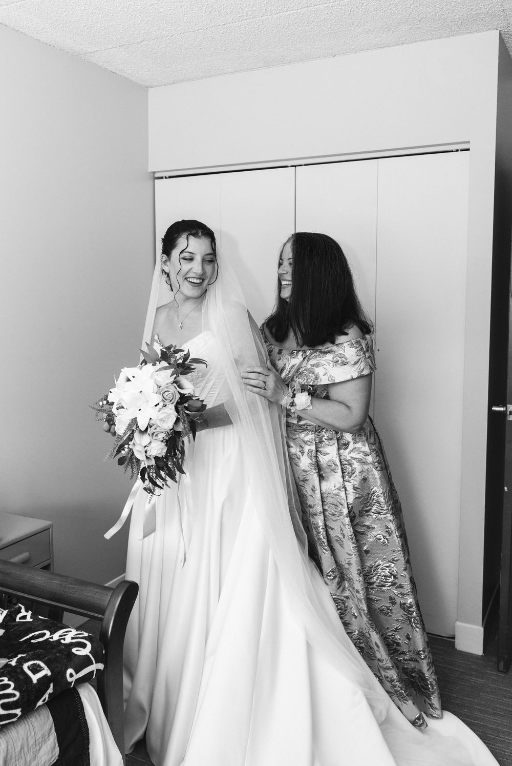 Smiling bride holding bouquet in white gown and veil assisted by woman in floral dress in black and white photo.
