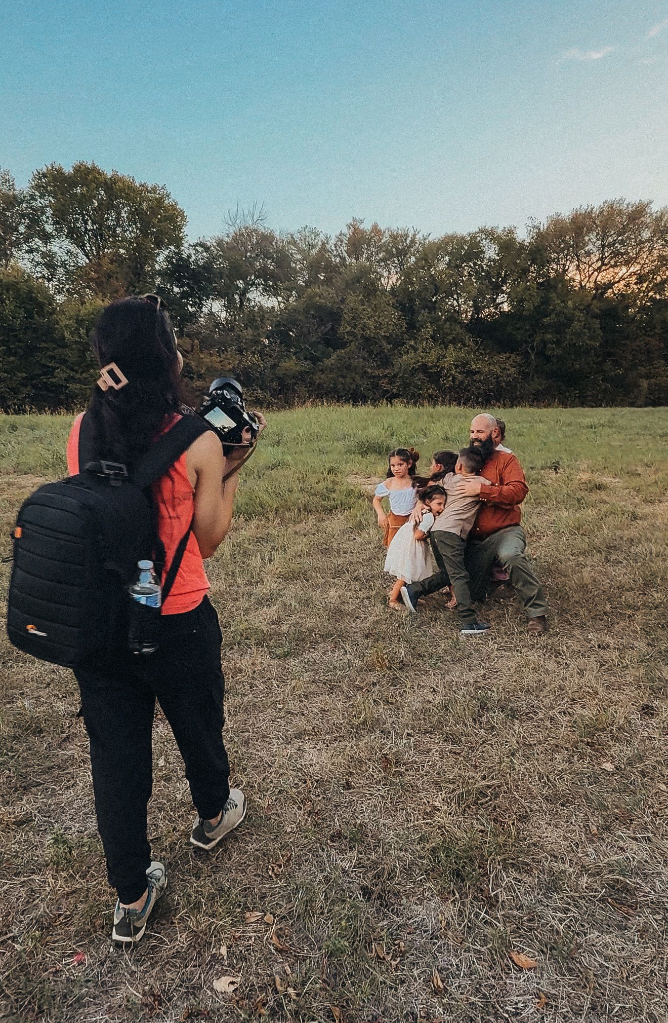 Woman in orange vest kneels in a field, taking a selfie with a drink, trees and vehicles visible in background.