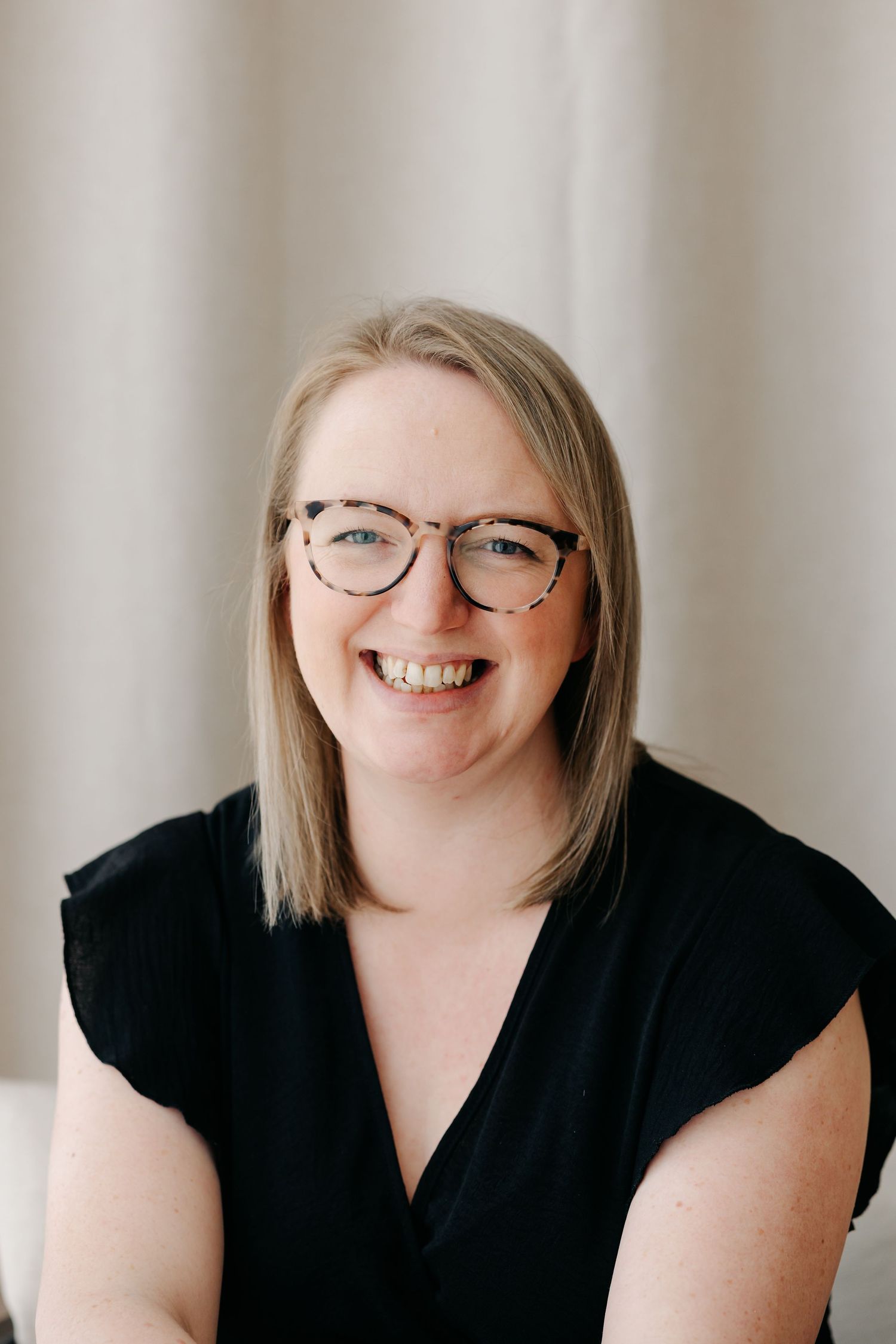 Smiling blonde woman with tortoiseshell glasses wearing a black sleeveless top, posed against a neutral background.