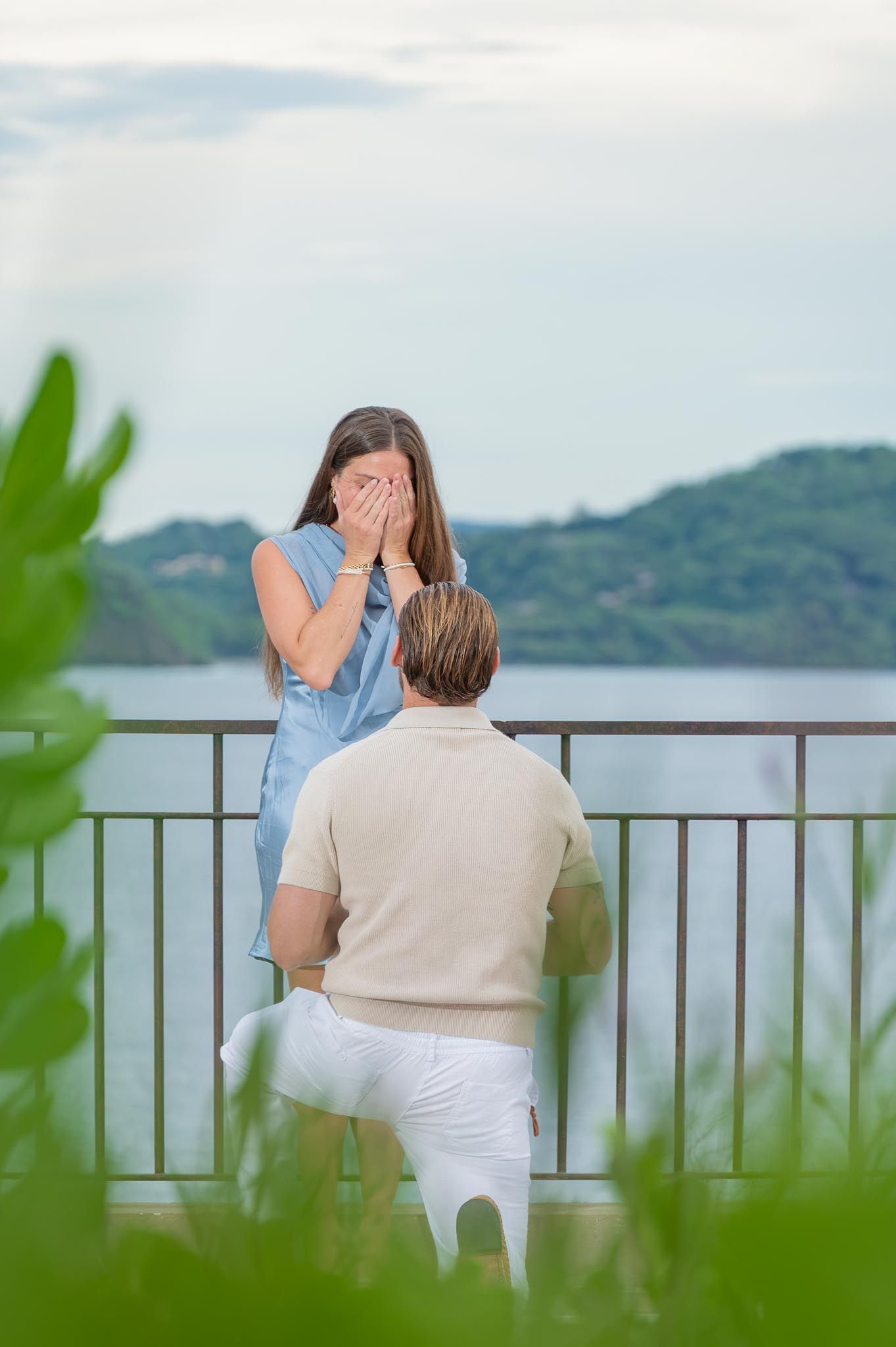 Smiling couple at dusk holding their hands together to show off a ring, surrounded by green trees and a blue evening sky.