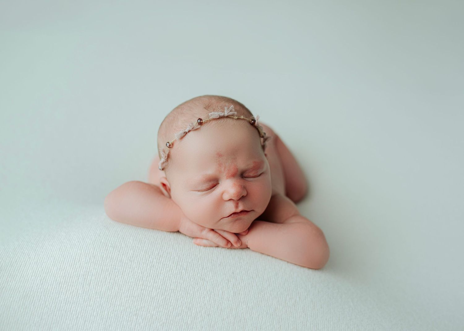 Peaceful newborn baby girl sleeping on white blanket wearing delicate floral headband in professional studio portrait.