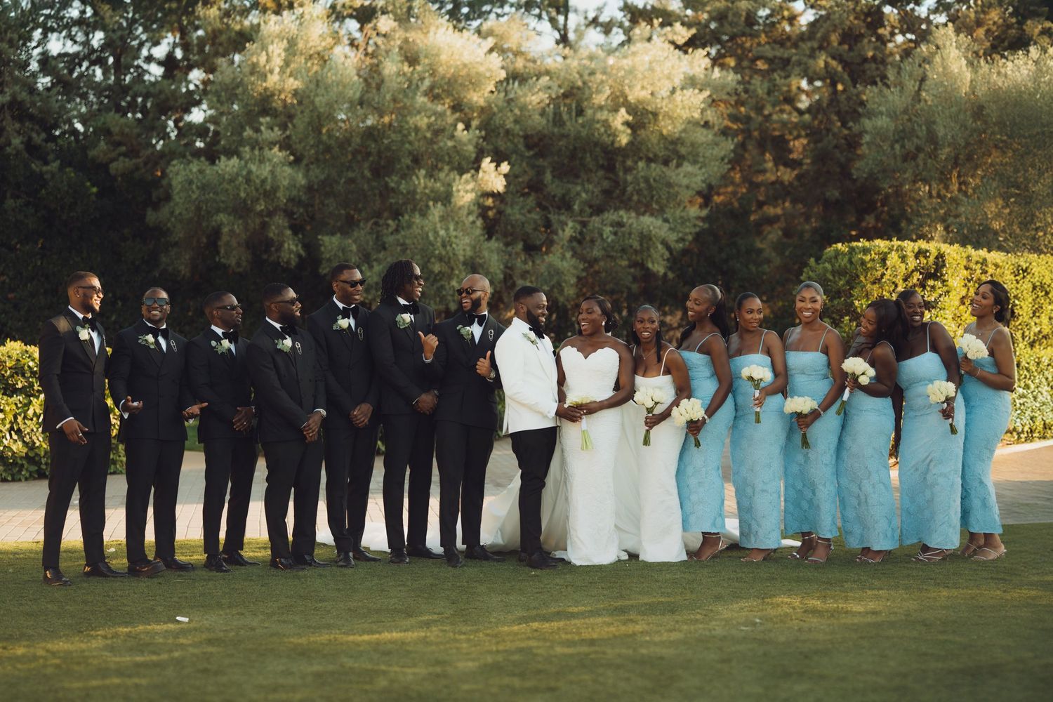 Wedding party in tuxedos and light blue bridesmaid dresses poses outdoors on green lawn with trees in background.