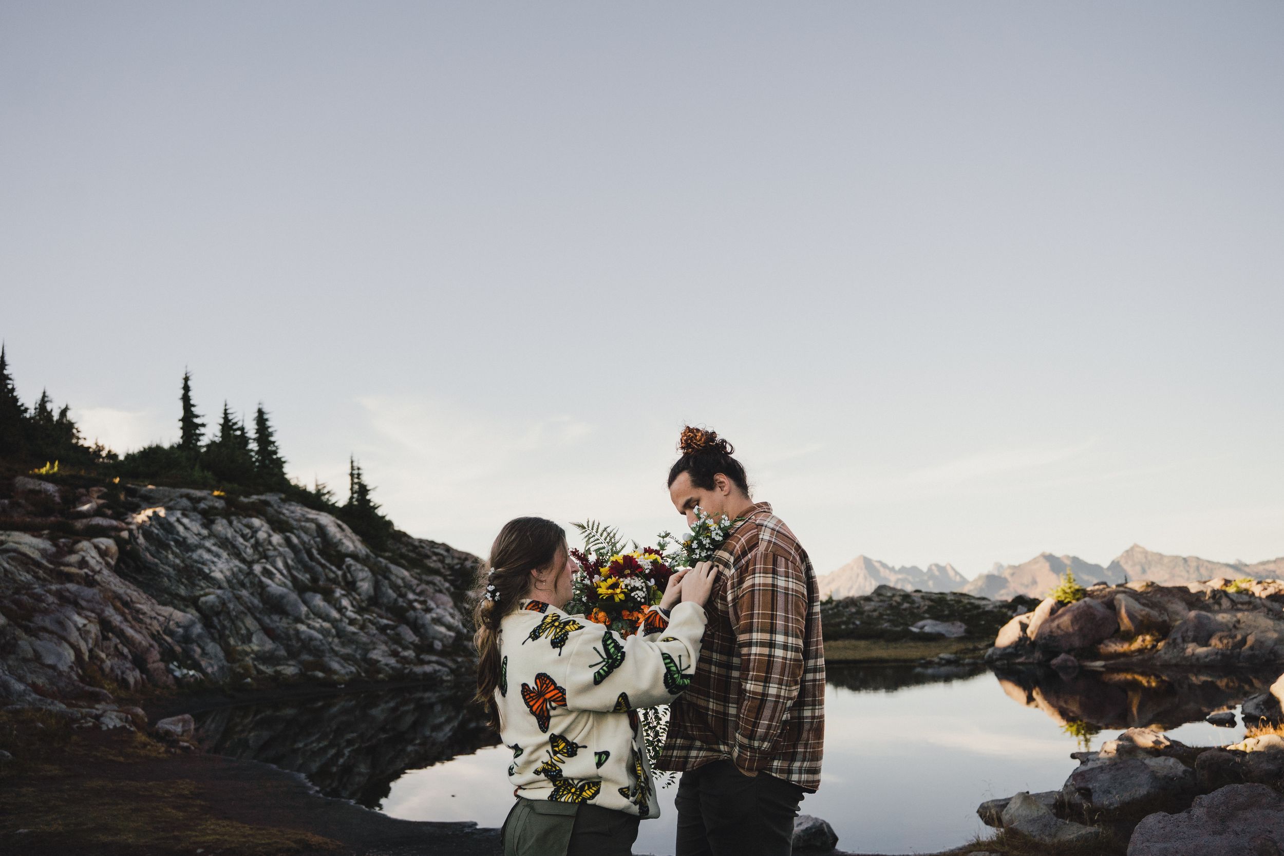 Couple holding a colorful bouquet beside a calm alpine lake with rocky shores, evergreen trees, and distant mountains at sunset.
