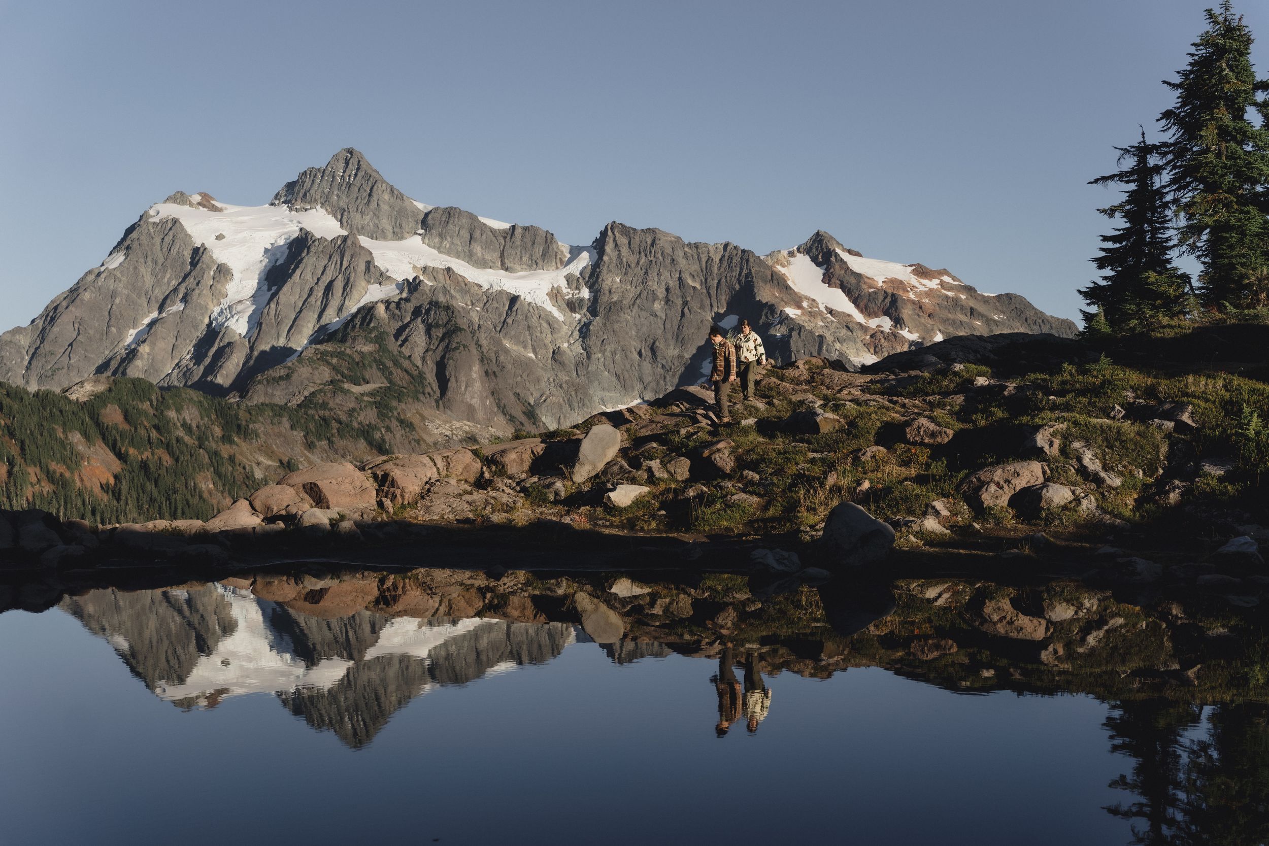 Two hikers stand on a rocky shore beside a calm alpine lake reflecting snow-capped mountain peaks under a clear blue sky.