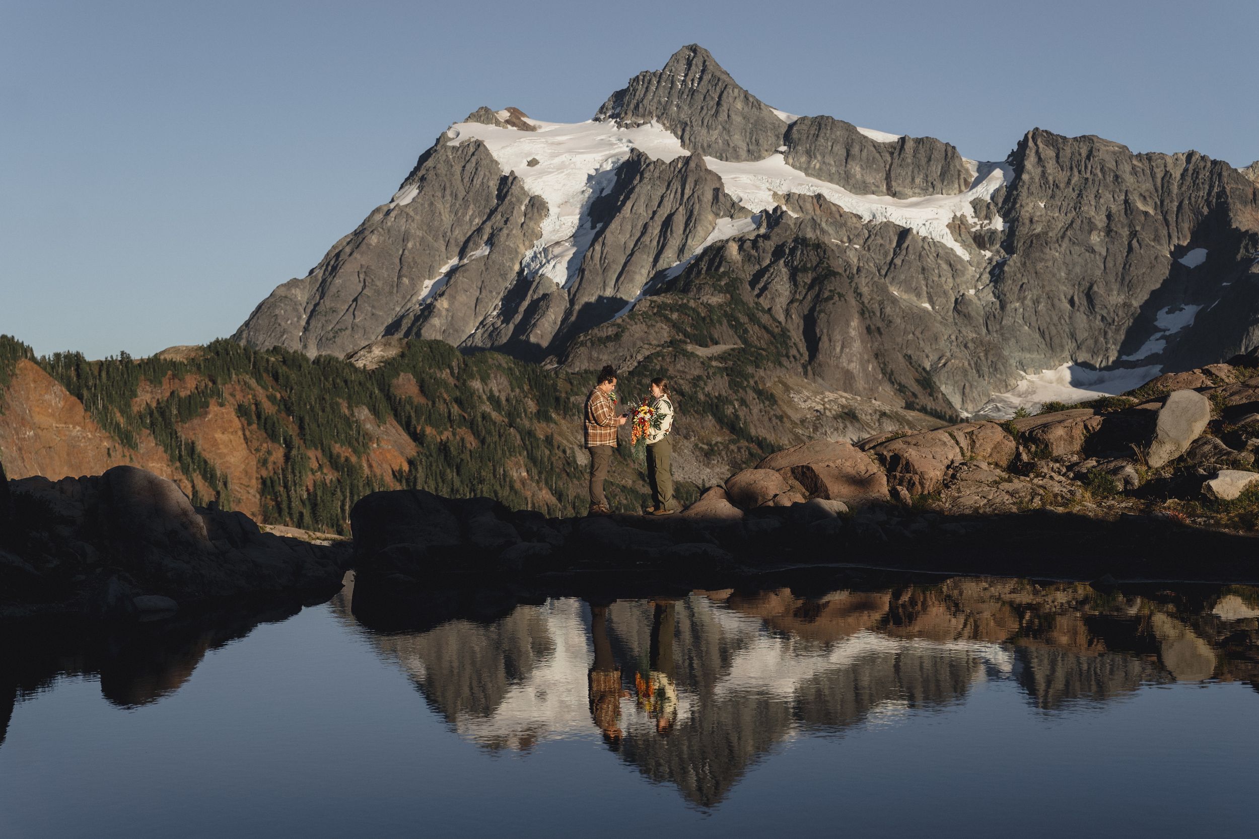 Two hikers stand on a rocky shore beside a calm alpine lake reflecting a snow-capped mountain peak under a clear blue sky.