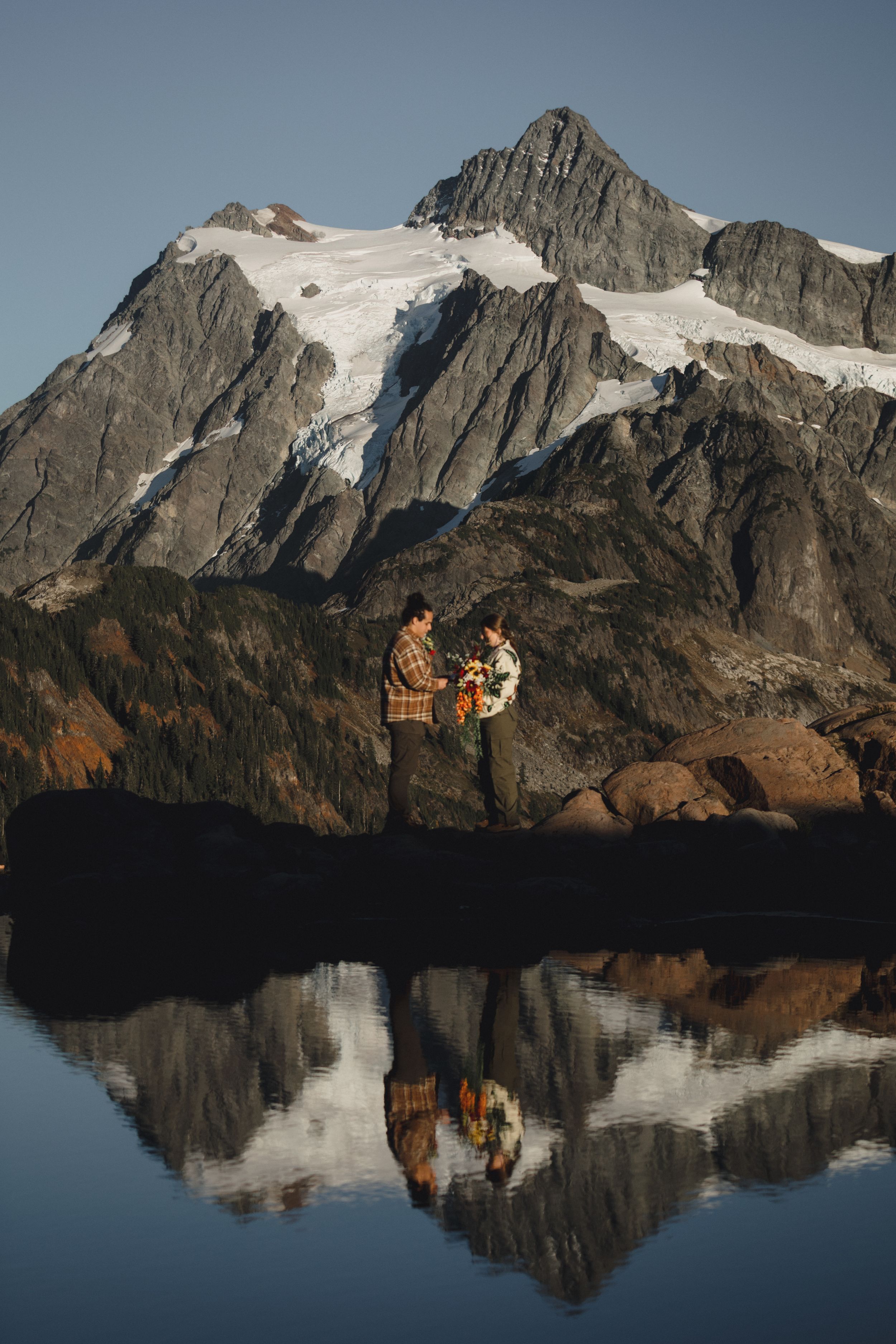 Couple in plaid jackets holding a bouquet on a rocky shore at sunset, with a snow-capped mountain reflected in a calm alpine lake.