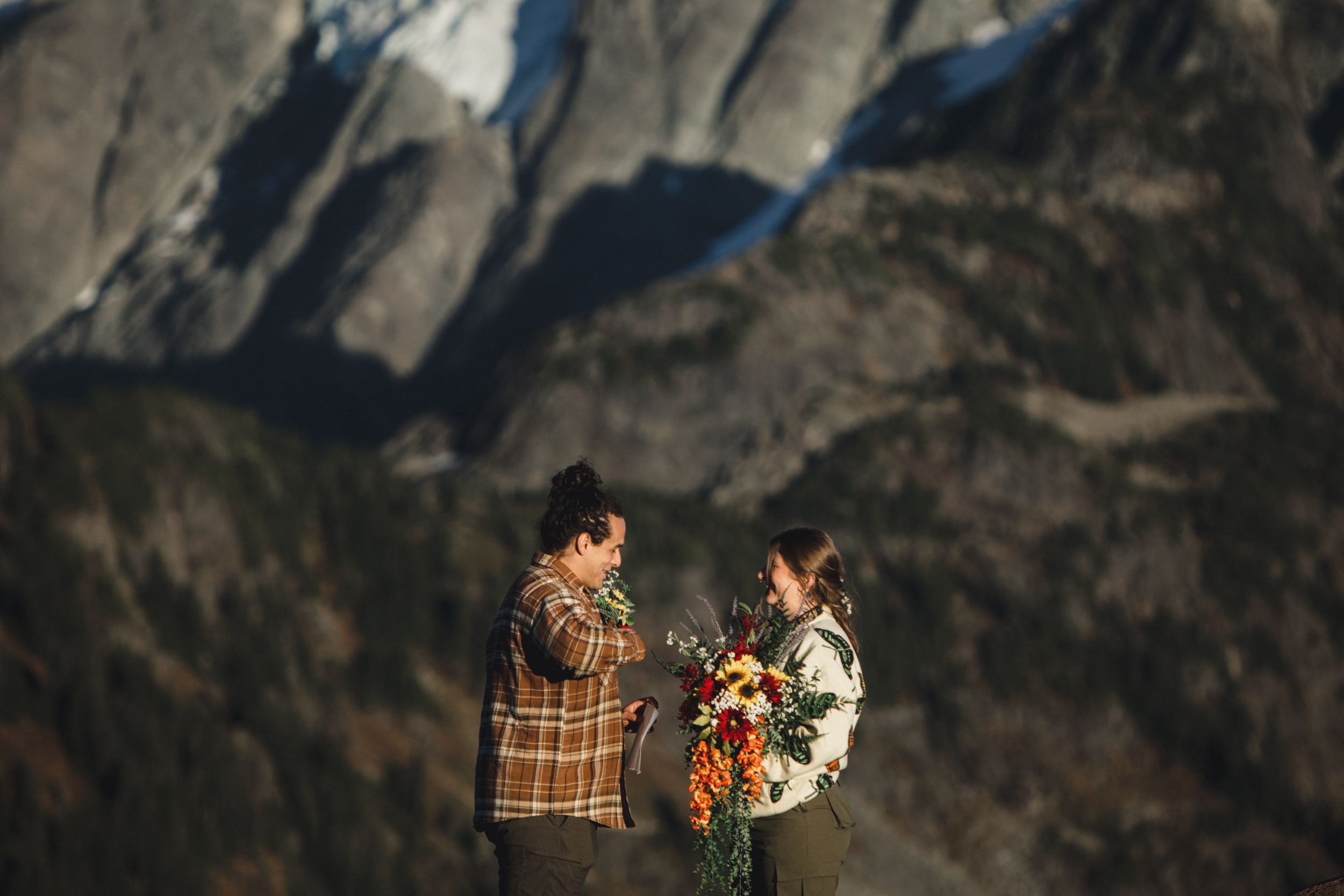 Couple in flannel jackets hold a colorful bouquet while embracing on a rugged mountain overlook during a golden hour elopement.