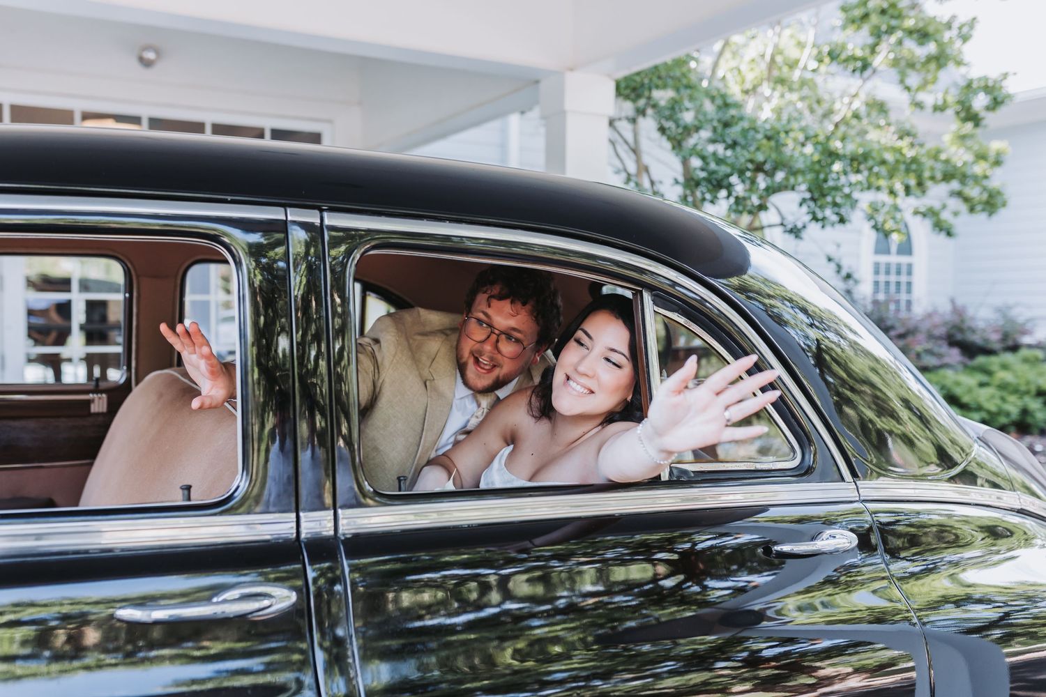 Smiling couple waving joyfully from the back window of a vintage black car, celebrating their wedding day.