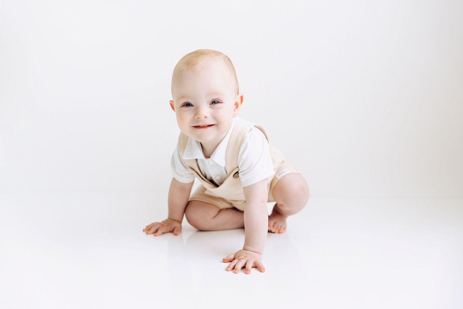 Happy baby boy in cream suspender onesie crawling and smiling on white studio floor.