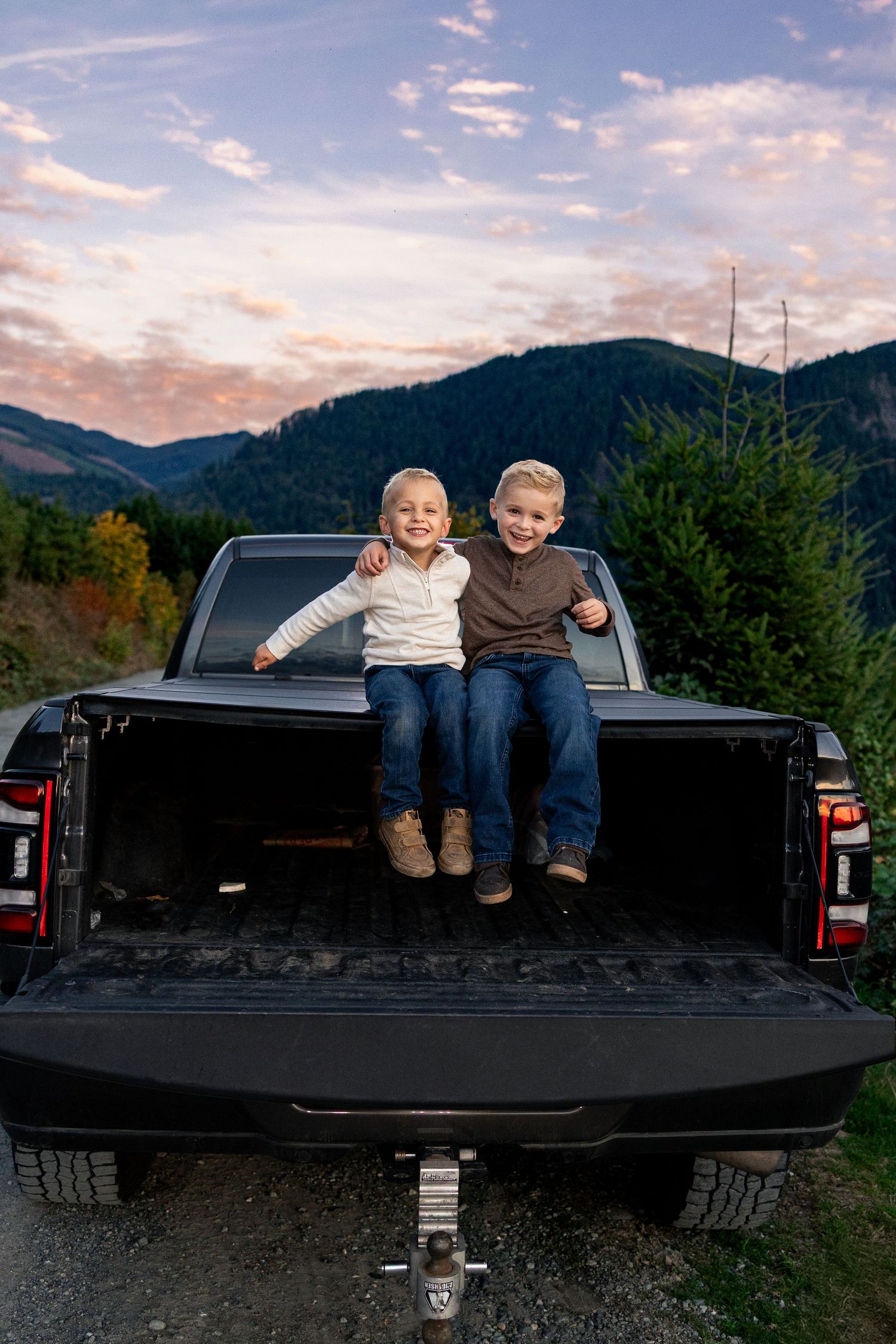 Two smiling children sit on a truck bed with mountain and sunset backdrop.