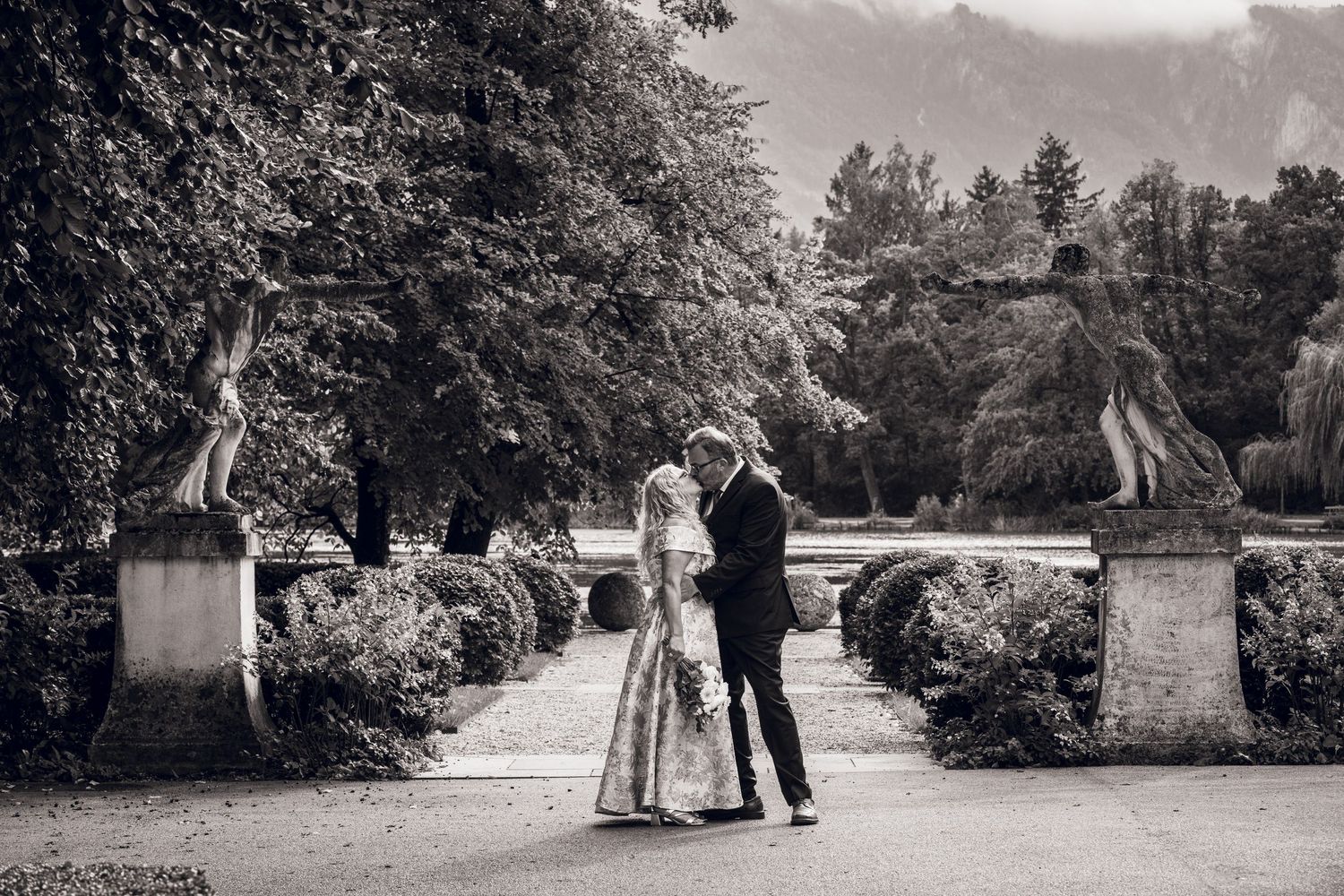 Bride in a white gown holding a bouquet embraces a groom in a navy suit on a garden path lined with statues, hedges and lush green trees.