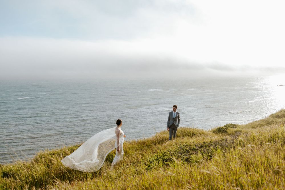 bride walking towards groom on clifftop field while pearl covered wedding cape flows in the wind on their san francisco elopement day