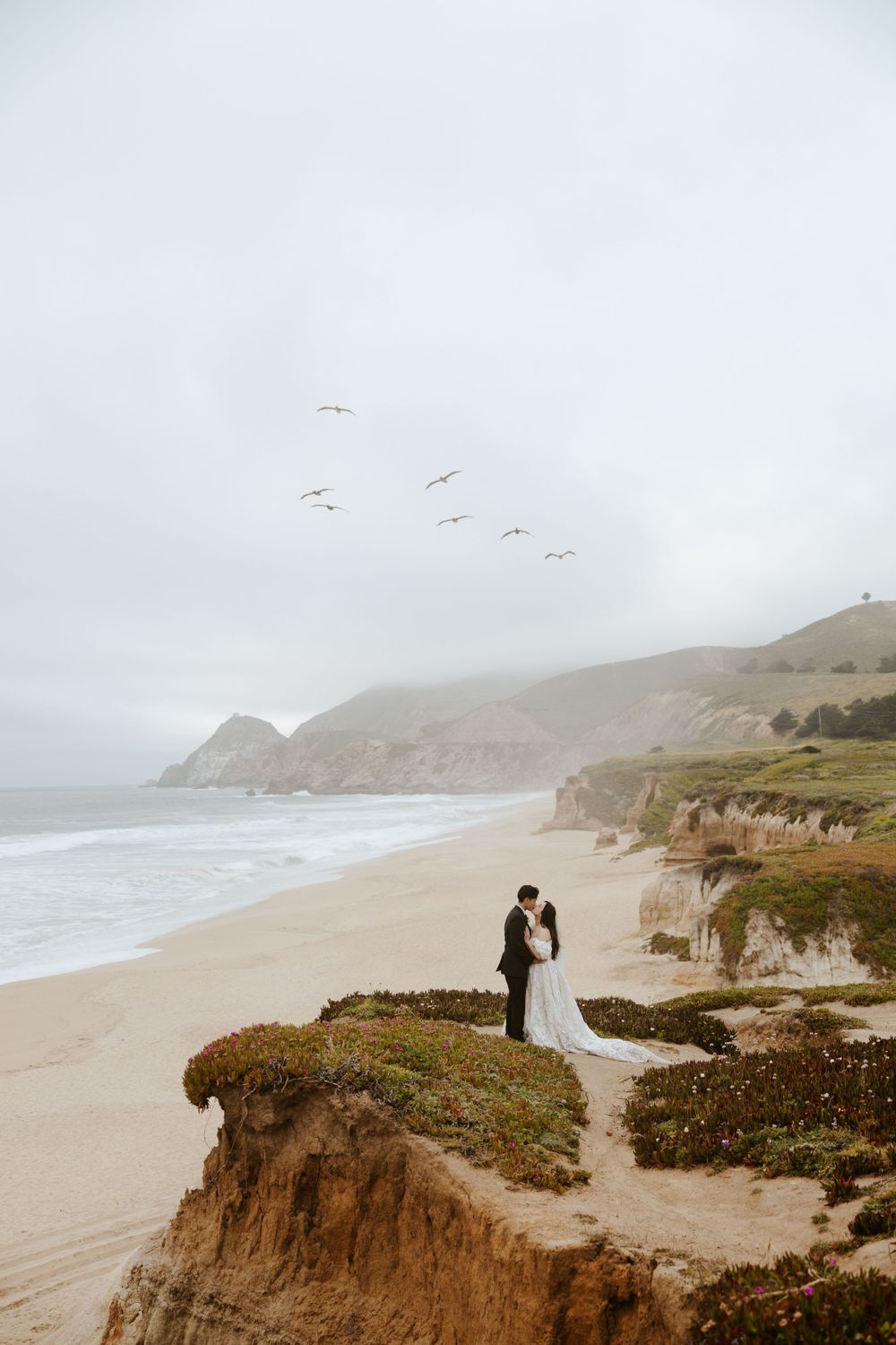 bride & groom kissing on a clifftop with ocean, beach, and mountains behind them while birds fly in the sky on their san francisco elopement