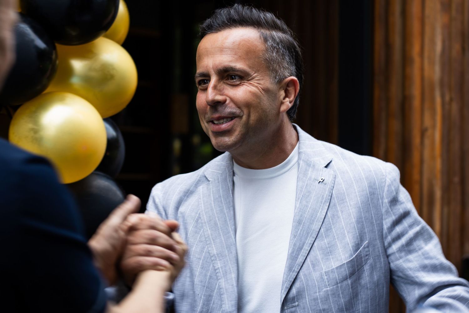 A smiling man in a grey blazer and white shirt at a party with black and gold balloons in the background.