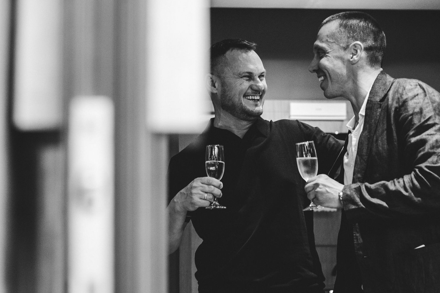 Two men laughing and toasting with champagne glasses at a formal event in black and white.