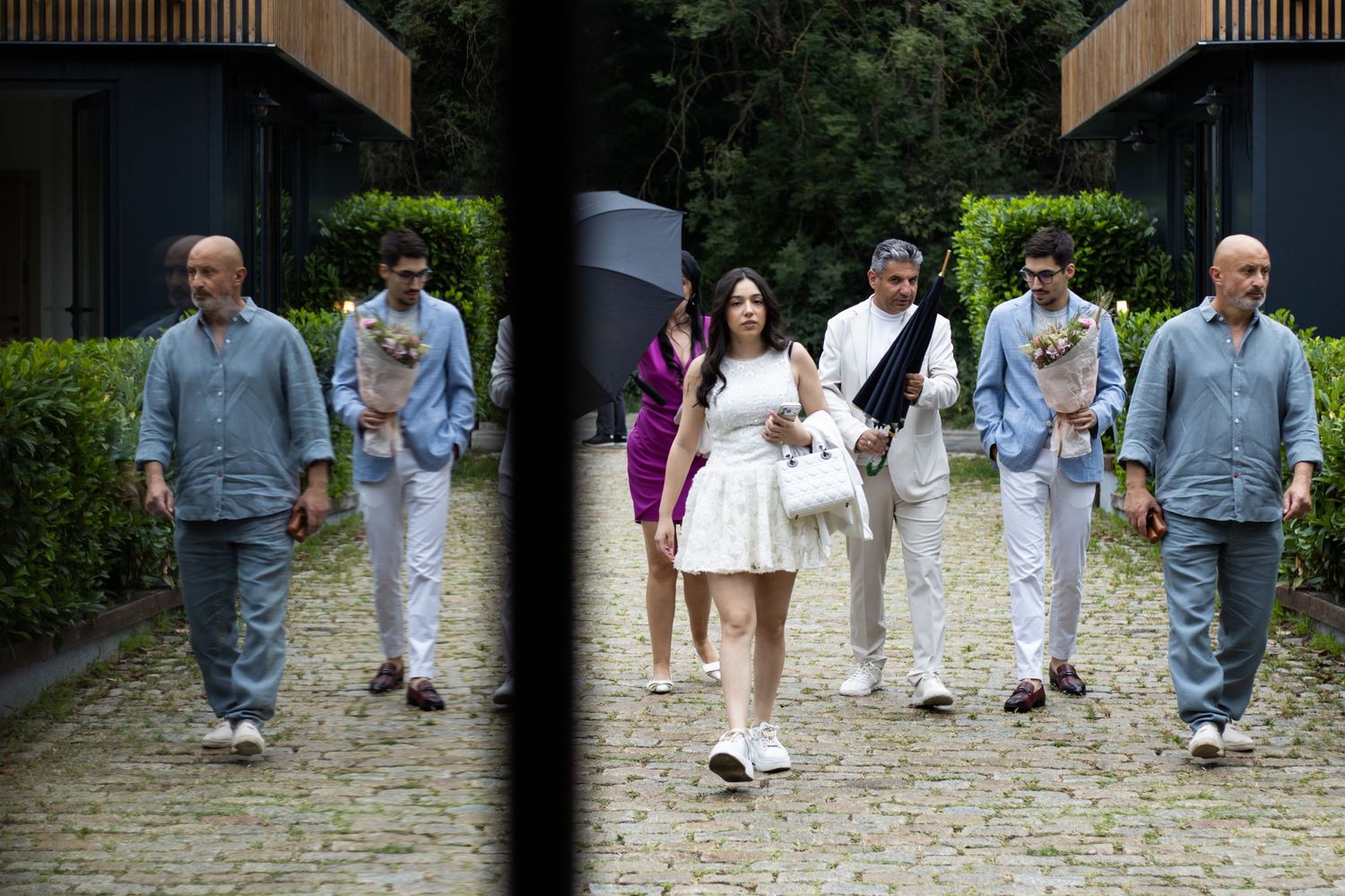 Group of elegantly dressed people walking together outdoors on a cobblestone path surrounded by greenery.