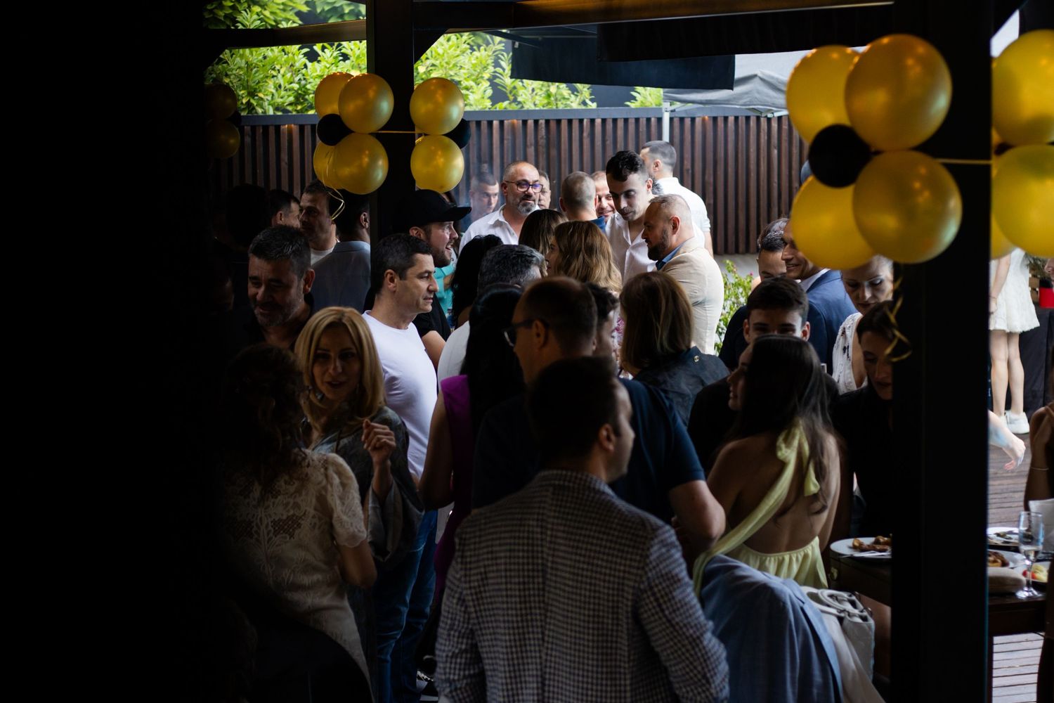 Crowd of people socializing at an outdoor party with yellow and black balloon decorations.