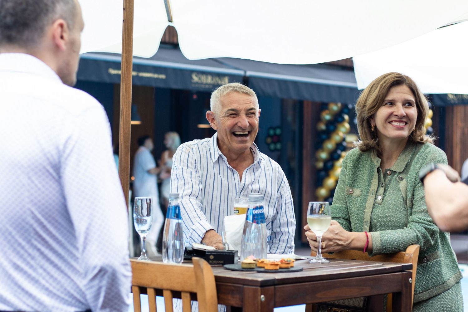 Three adults laughing and socializing outdoors at a sunny event, enjoying wine and snacks at a wooden table.