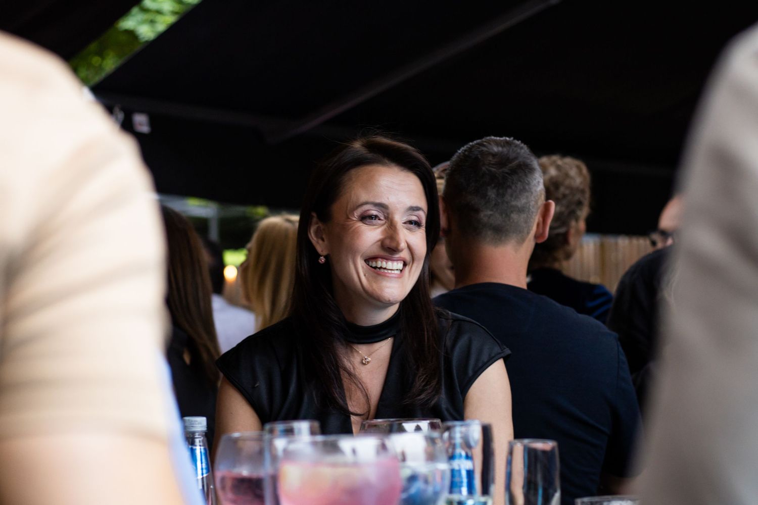A smiling woman in a black top and choker necklace enjoys a social gathering at an outdoor dining event.