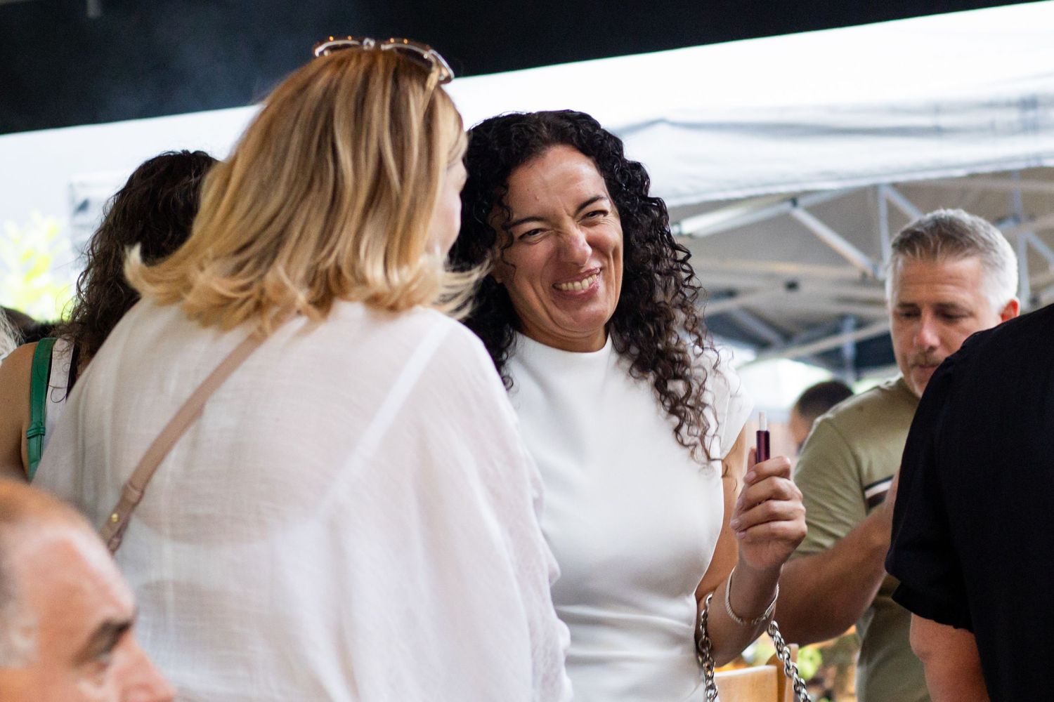 Two women smiling and chatting at an outdoor social event, one holding a phone, dressed in white outfits.