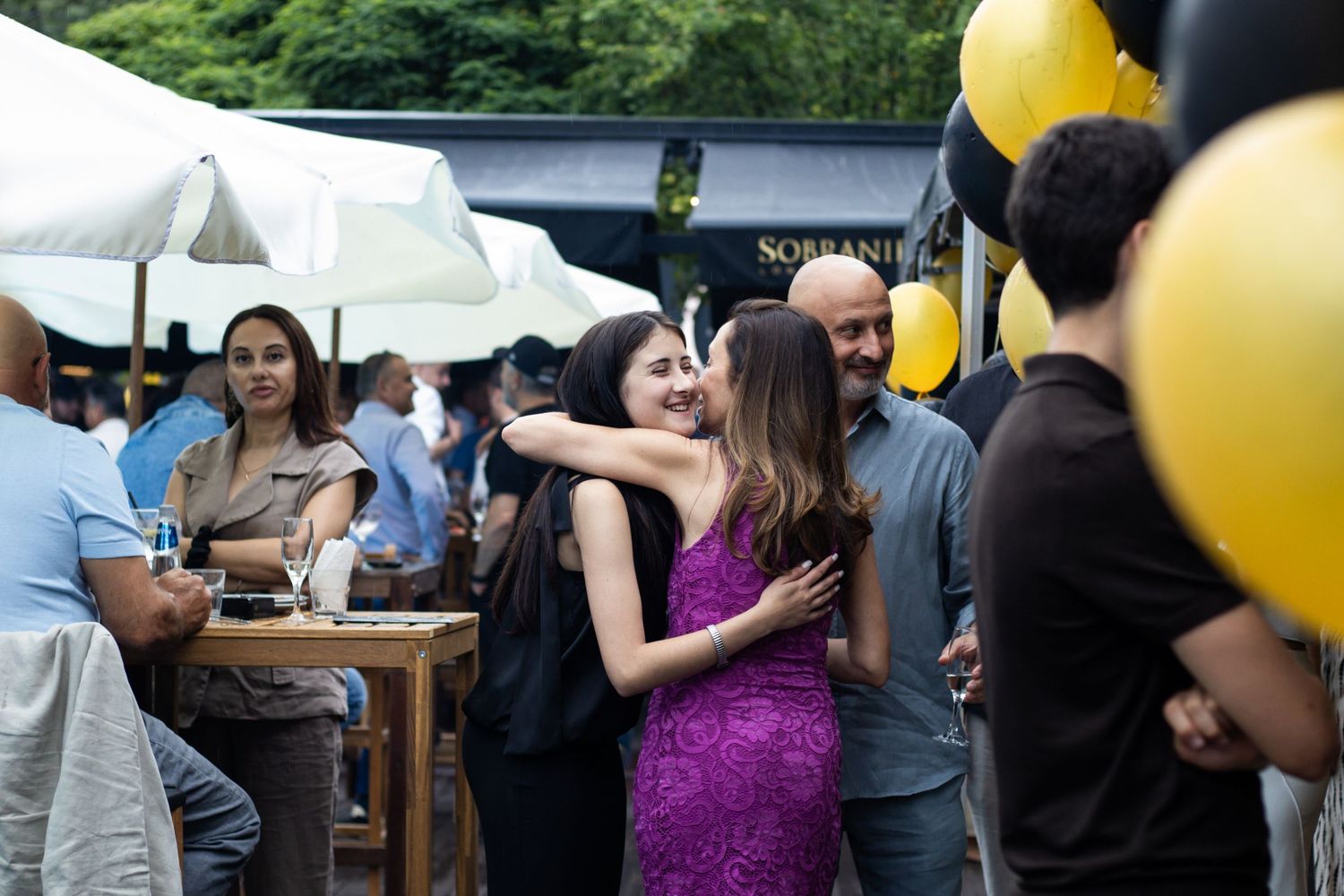 Two women embrace warmly at an outdoor party with black and gold balloons at a restaurant patio.