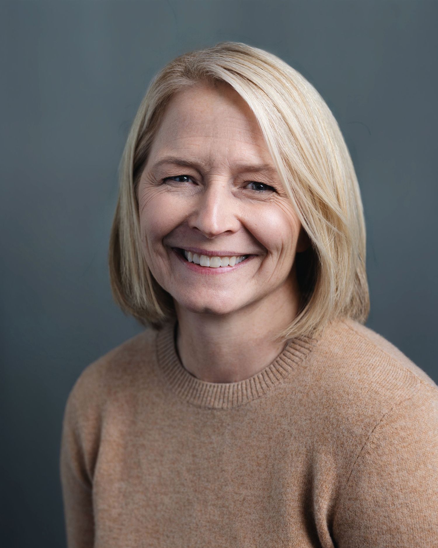 Professional headshot of a smiling blonde woman in a beige sweater against a gray background.