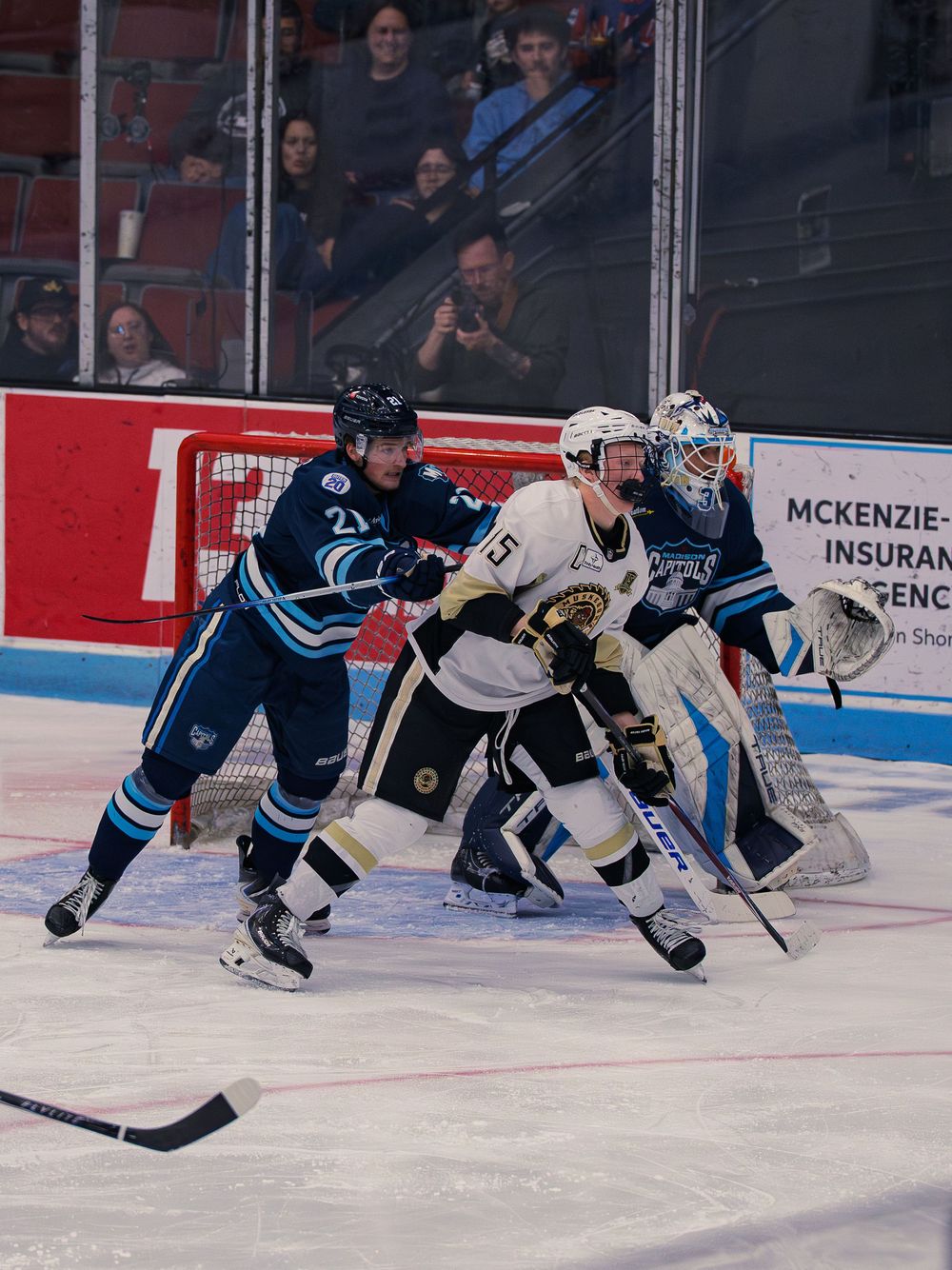 A Capitols player number 21 ties up a white Lumberjacks player number 15 in front of the Capitols goaltender guarding his crease.
