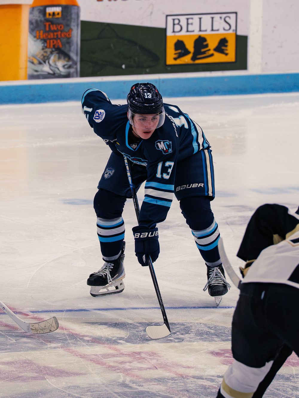 A Madison Capitols player number 13 crouches low over his stick in a ready stance, facing off against an opponent at the edge of frame.
