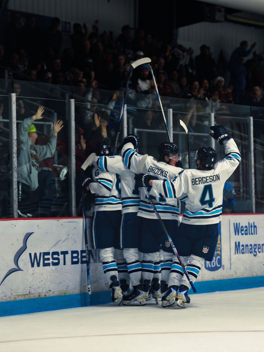 A group of Madison Capitols players huddle together to celebrate a goal along the boards as fans cheer from the stands behind.